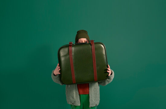 Woman in fur coat holding green suitcase covering face against green background