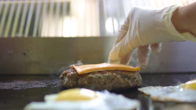 Chef putting slice of orange cheese on hot grilled beef burger patty with fried eggs on commercial griddle in restaurant kitchen