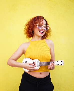 Red haired woman in yellow top playing ukulele joyfully against wall