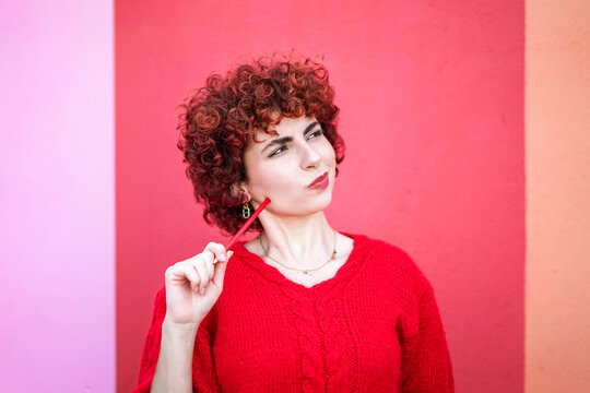 Curly redhead in red sweater thinking with pencil against vibrant background