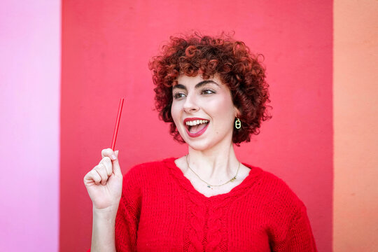 Redhead with curly hair smiling and holding pencil against colorful wall