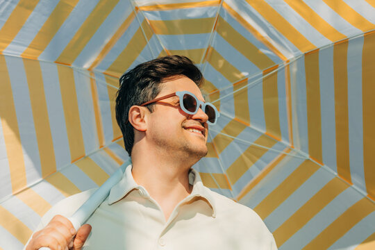 Smiling man with sunglasses under striped summer umbrella outdoors