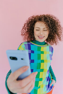Smiling woman with curly hair taking selfie on phone against pink background