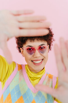Smiling woman wearing heart shaped glasses framing face on pink background