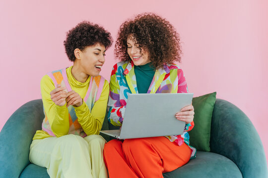 Two friends with curly hair using laptop and smiling on sofa indoors