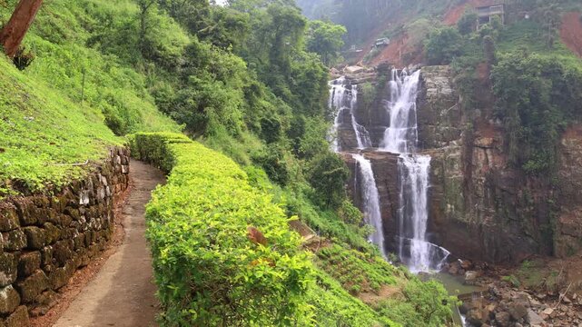 Ramboda Waterfall near Nuwara Eliya town in Sri Lanka. Majestic multi-tiered waterfall cascading down a lush green mountain slope with a stone path and vibrant foliage. Slow motion footage.