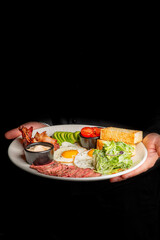 Hands holding a white plate with a keto-style breakfast: fried eggs, sliced steak, bacon, avocado,...
