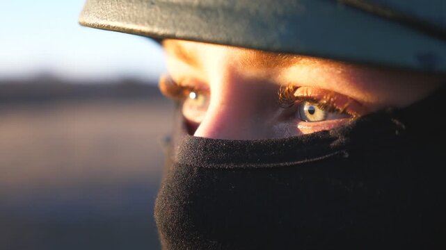 Close up green eyes of female ukrainian army soldier looking at sunset against blue-yellow flag. Sad sight of military woman in helmet and balaclava at countryside. Resistance to russian invasion