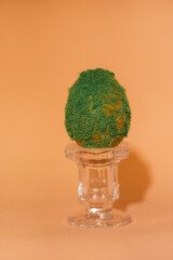 Decorative green Easter egg displayed on a clear glass pedestal against a soft orange background,...