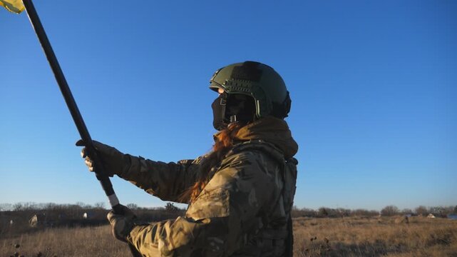 Dolly shot of young girl in military uniform waving flag of Ukraine against background of sunset. Female ukrainian army soldier lifting national banner on the field. Victory against russian aggression