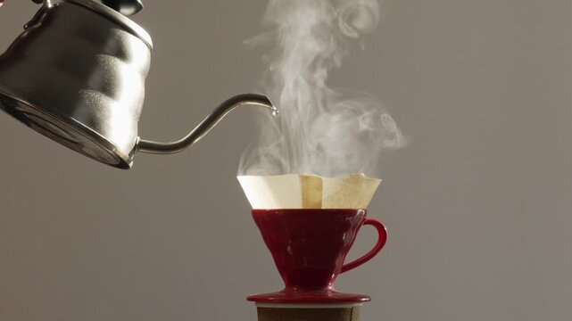 Barista pouring boiling water from silver gooseneck kettle into red ceramic coffee dripper with filter