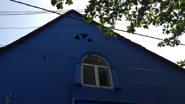 In Kochi&rsquo;s Jew Town, this vibrant blue building features a steeply pitched roof, arched window, & striking exterior wall. Set against lush greenery & power lines, its bold facade Kerala's streetscape.