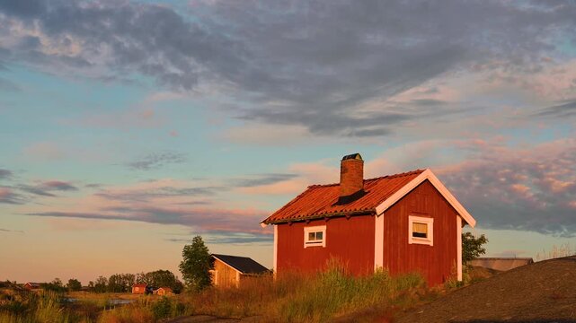 Red wooden cottage in Swedish archipelago during colorful sunset with moving clouds and golden hour lighting