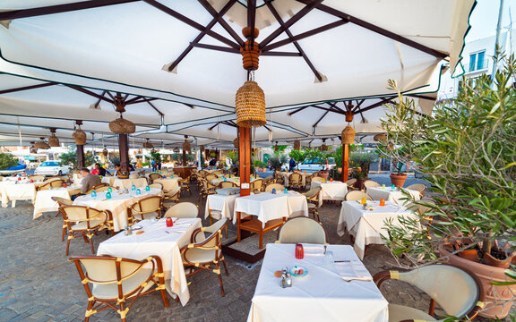 Outdoor restaurant with tables and chairs under canopy umbrellas in Capri, Italy. Tables covered with white cloth and colorful glass holders. Rustic lamps and potted plants create cozy atmosphere