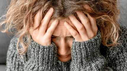 Fototapeta premium Woman with curly hair wearing a cozy sweater is holding her head in distress, expressing feelings of anxiety and emotional struggle in a soft-lit indoor environment
