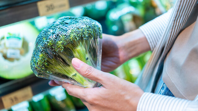 Close-up of a shopper's hand holding fresh broccoli wrapped in plastic film on a supermarket shelf. Choosing healthy green vegetables for nutrition during grocery shopping.