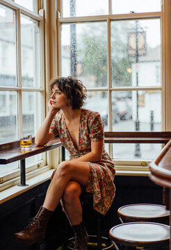 Nachdenkliche junge Frau im Blumenkleid an einem verregneten Fenster in einem traditionellen Pub