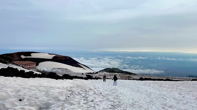 Timelapse of family descending snowy slope near Silvestri crater on Mount Etna. People moving across volcanic landscape with Rifugio Sapienza in background under dramatic sky in early spring.