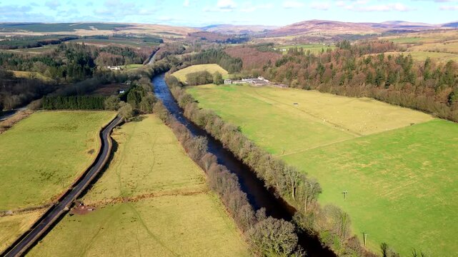 The River Esk winds through green fields and dense trees at Gilnockie Tower in the Scottish Borders. This scene shows a clear view of the landscape near Langholm on the A7. Hills rise in the backgroun