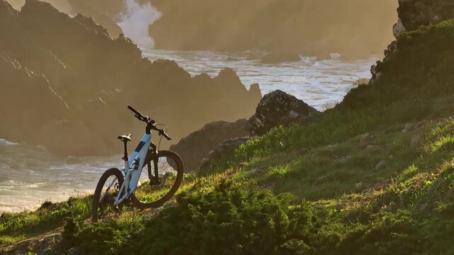 Electric adventure bike parked on grassy cliff over Atlantic ocean and dramatic rock formations, illuminated by the golden light of setting sun in Portugal. Active sports, recreation, outdoor activity