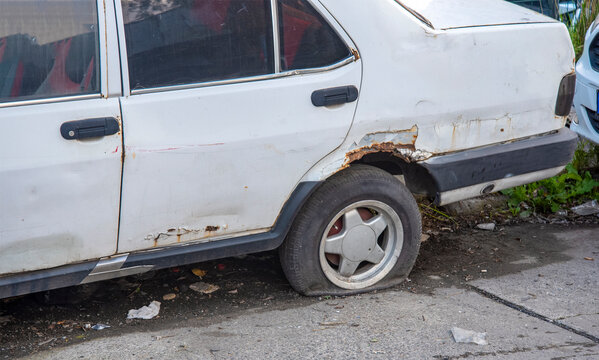 Old white car parked on the street with a flat tire and rust damage in a city setting during daytime
