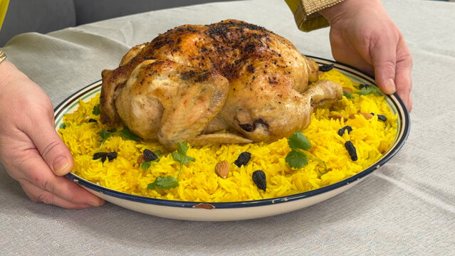 Hands serving a whole roasted chicken on a platter of yellow rice with nuts and raisins.