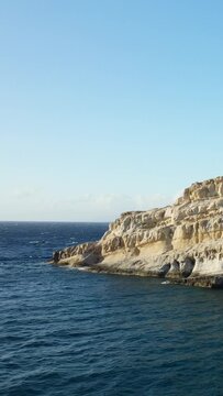 Rocky limestone cliffs and caves rise above the blue waters of the Mediterranean Sea at Matala, Crete, under a clear sky. The dramatic coastline is bathed in natural sunlight, highlighting the unique