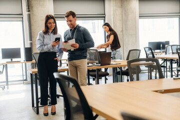 Two professionals discussing work while using mobile devices in a modern office space with desks and computers arranged in the background