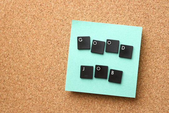 Black keyboard keys spelling "GOOD JOB" on a light blue sticky note pinned to a cork bulletin board.
