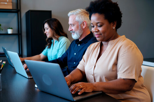 Middle aged Black woman, middle aged Caucasian man, and young adult Caucasian woman working on laptops in modern office setting, all focused on screens and typing together