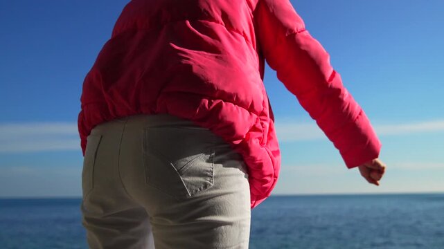 Ocean throwing woman in bright pink jacket stands by the sea on a sunny day and tosses objects into the blue water