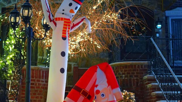 Funny inflated snowman and Santa wave outdoors under the falling snow. Light garlands are shining in the backdrop. Christmas in USA.