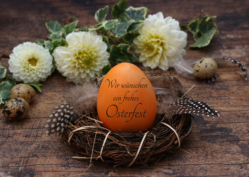 Orangfarbenes Ei  mit dem Schriftzug wir w&uuml;nschen ein frohes Osterfest in einem Nest aus Zweigen und Federn.