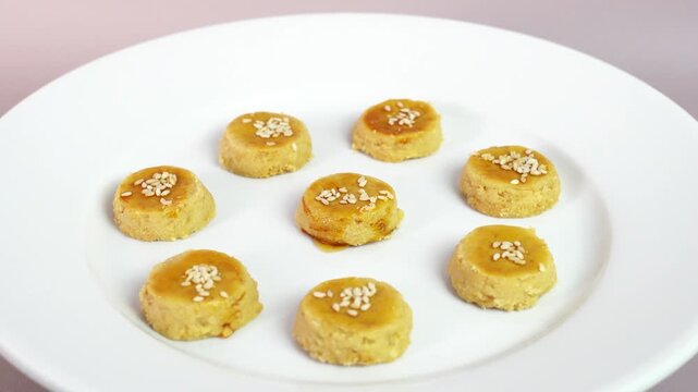 Golden bite-sized cookies topped with sesame seeds arranged on a white plate against a soft pastel background. Homemade bakery snack ideal for tea time