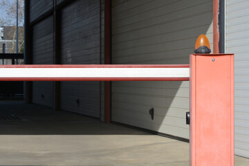 Fototapeta premium A red and white boom barrier gate with an orange warning light stands in front of closed industrial garage doors