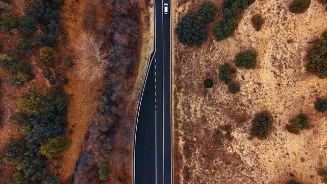 Aerial top down view of car on asphalt road in Spain. High angle shot of white vehicle driving on two lane highway through arid countryside in Spain.