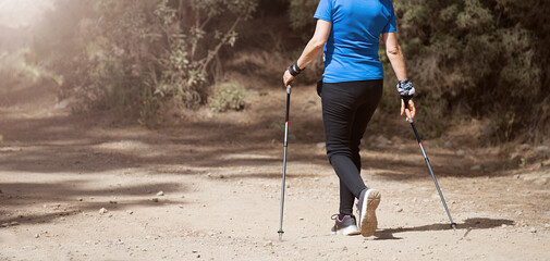 Senior woman walking in forest with Nordic walking sticks, sunny summer day. An active lifestyle in retirement, a hike in nature