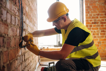 Male electrician wearing yellow safety vest and hard hat works on electrical wiring against a brick wall in a construction site with natural light coming through a window