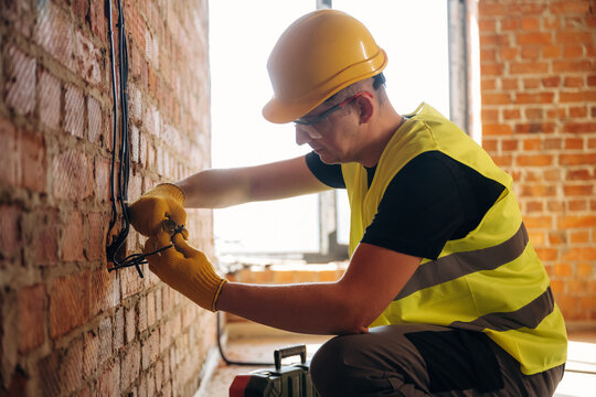 Male electrician wearing yellow safety vest and hard hat works on electrical wiring against a brick wall in a construction site with natural light coming through a window