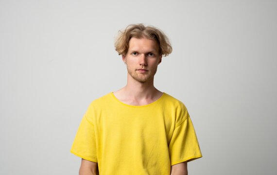 Young man with blonde hair and a beard wearing a yellow t-shirt, standing and looking straight ahead, with a serious expression, isolated on a white background
