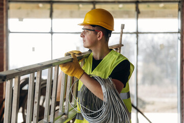 Male construction worker wearing yellow hard hat and safety vest carries ladder while holding electrical wires in a partially constructed building with large windows © standret