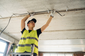 Male electrician in safety vest installing electrical wiring on ceiling while standing on a ladder inside a modern building under construction with large windows