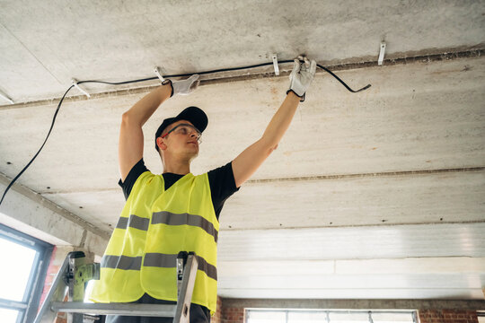 Male electrician in safety vest installing electrical wiring on ceiling while standing on a ladder inside a modern building under construction with large windows