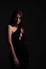 Naklejka premium Confident woman in a one-shoulder black evening dress with keyhole cutout, posing against a dark background; low-key lighting adds mystery and elegant, sophisticated glamour