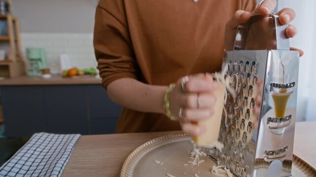 Tilt down closeup shot of Caucasian teen girl grating cheese over plate in bright kitchen interior, showing food preparation and everyday cooking routine