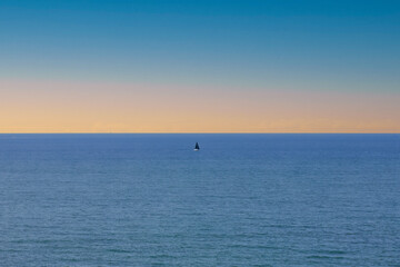 Obraz premium Minimal seascape with a single sailboat on the horizon line in calm blue water under a soft pastel sky.