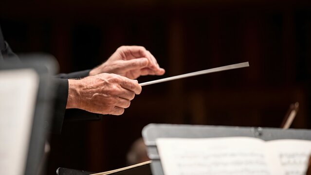 A conductor's hands hold a baton while leading an orchestra on stage
