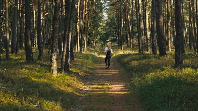 Blonde woman walks along a wide forest path, makes a 360 turn to look around, then continues walking. Peaceful nature scene with calm atmosphere and exploration vibe