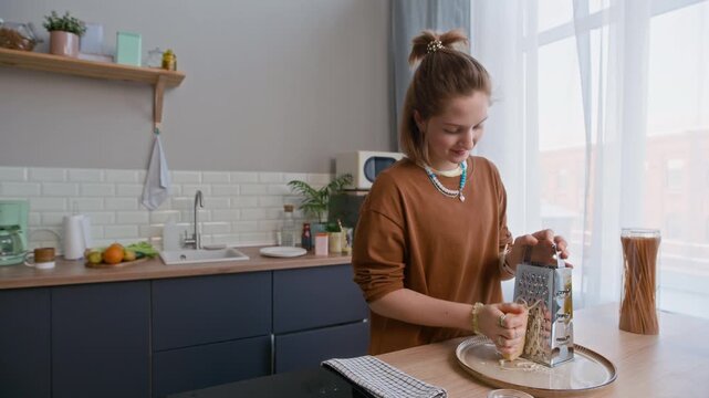 Medium eye level shot of Caucasian teenage girl grating cheese while cooking spaghetti in bright modern kitchen, showing pasta preparation and home cooking process