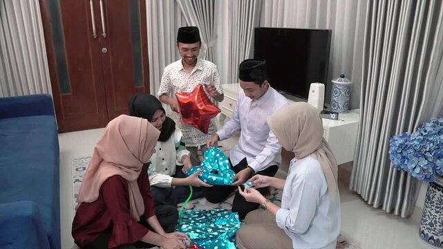 Asian muslim family unwrapping eid gifts in cozy living room with turquoise star balloons, parents and three daughters wearing